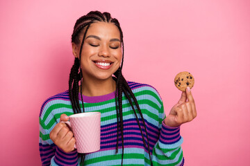 Smiling young woman wearing a colorful striped sweater holding a cookie and coffee cup on a pink...
