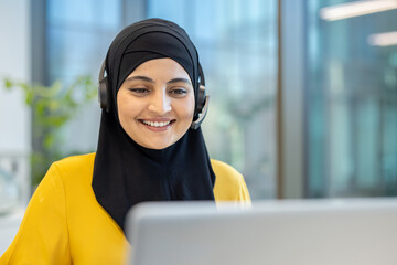 Muslim woman in a hijab smiling with headset while providing customer service and working on laptop in a modern office, remote support agent offering friendly assistance