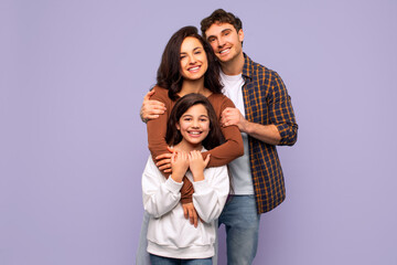 Portrait of happy European dad and mommy embracing their daughter, standing on purple studio background. Healthy family relationship concept