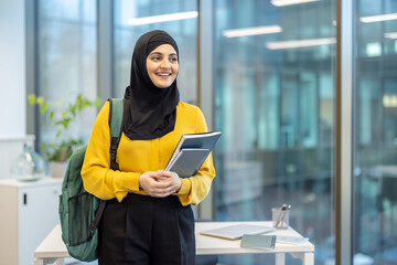 Young muslim woman smiling, carrying a backpack and holding notebooks, standing in a modern office or university corridor, representing education, career, and diverse workforce