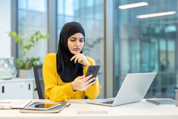 Muslim businesswoman wearing a hijab sitting at her modern office desk, reading her smartphone with a concerned and thoughtful expression while working on her laptop
