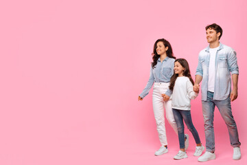 Fototapeta premium Family of three holding hands and walking over pink background, parents and their daughter in casual clothes, full length, free space