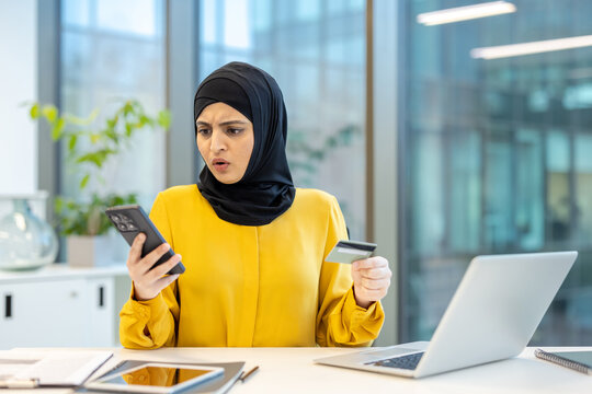 Muslim businesswoman wearing a hijab expressing shock and frustration while looking at her smartphone and holding a credit card, experiencing an online shopping payment error or fraud