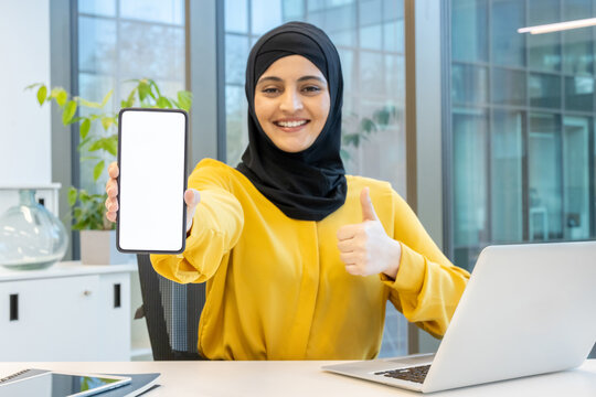 Muslim businesswoman in hijab at office desk, smiling and giving thumbs up while presenting a smartphone app on a blank screen, confident and professional endorsement - Powered by Adobe
