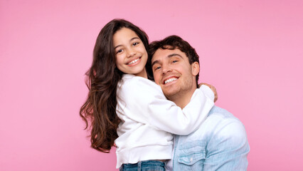 Fototapeta premium Portrait of happy European daughter embracing her father, man holding preteen girl in arms, both smiling at camera over pink studio background