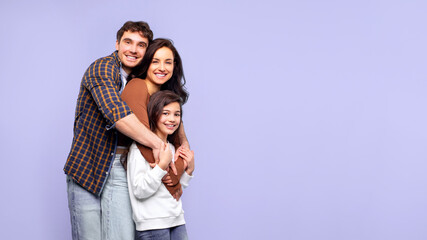 Fototapeta premium Family portrait. Happy loving father, mother and their daughter posing isolated on purple studio background, hugging each other and smiling at camera, panorama with free space