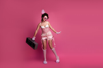 A young woman in a pink corset poses playfully against a solid pink background. She holds a vintage boombox
