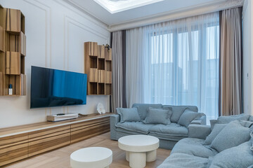 A spacious living room features a large blue sofa, stylish coffee tables, and wooden shelving. Sunlight streams through floor-to-ceiling windows, enhancing the modern atmosphere.