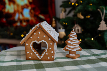 Handmade gingerbread house with heart-shaped door and Christmas tree cookie on checkered tablecloth with festive bokeh lights background. Holiday baking traditions, cozy celebration and sweet treats.