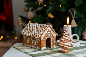 Decorated gingerbread house with detailed icing and Christmas tree cookie beside burning candle on festive table with bokeh lights. Holiday baking crafts, cozy winter celebration and traditions.