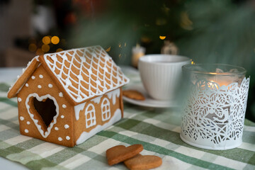 Cozy Christmas composition with homemade gingerbread house decorated with icing, burning candle in white holder, and cup of tea on checkered tablecloth. Winter holidays, warmth, and festive mood.