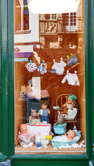Colorful display of dolls in shop window, featuring various outfits hanging, cozy setting with accessories, and playful elements, creating whimsical atmosphere for children's imagination and play