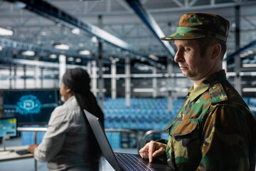 Military officer reviewing mission critical data on laptop in server room command center. Soldier wearing uniform analyzing satellite communications on notebook in army data center