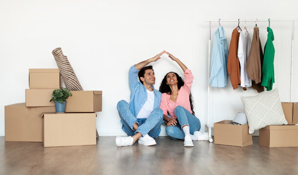 Mortgage for Young Families. Happy couple moving to their new own home, joining arms making symbolic house roof of hands above head, sitting on floor among cardboard boxes, smiling. Property Insurance