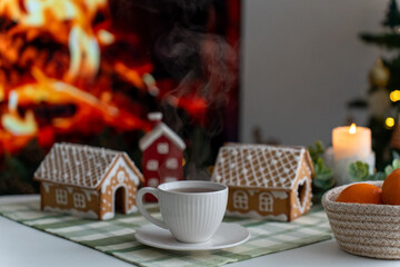Cup of hot tea with steam on table near gingerbread houses, burning fireplace and candle creating cozy Christmas atmosphere. Winter holidays, home comfort, festive decoration and warm family mood.