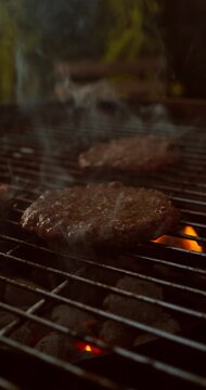 Vertical video of juicy burger patties sizzling on grill with fire and smoke. Appetizing barbecue cooking closeup 4k 60fps.
