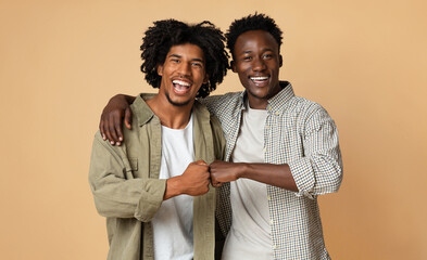 Portrait Of Two Cheerful Guys Embracing And Making Fist Bump Gesture, Happy Young African American Friends Having Fun Together While Standing Over Beige Studio Background, Copy Space