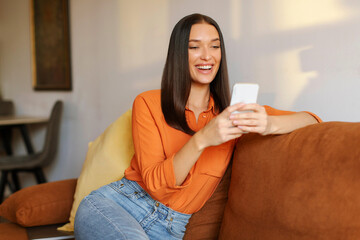Happy young woman relaxing on comfortable couch, holding smartphone, chatting in social networks,...
