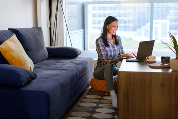 Young woman freelancer working on laptop, sitting on floor by coffee table in living room, having video call or surfing web, free space