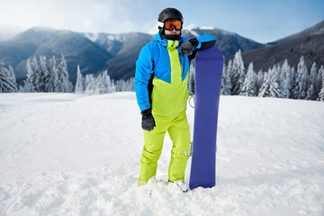 Skier stands proudly on snowy mountain slope, showcasing bright winter gear and snowboard against stunning backdrop of snow-capped peaks and pine trees © verona_studio