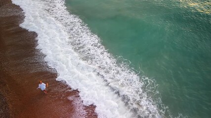 A middle-aged woman in a white dress and hat walks barefoot along the foamy shoreline, enjoying the sunrise and the calm beauty of the sea.