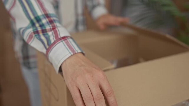 Man opens cardboard box with hand in building interior wearing bright plaid shirt; fresh start excitement.