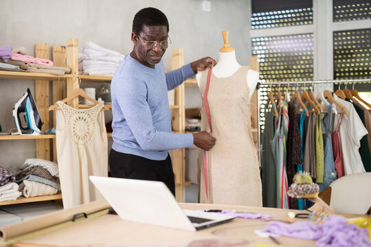 Professional fashion designer participating in remote consultation via video call using laptop in workshop, refining linen garment on mannequin while communicating with client or colleague..