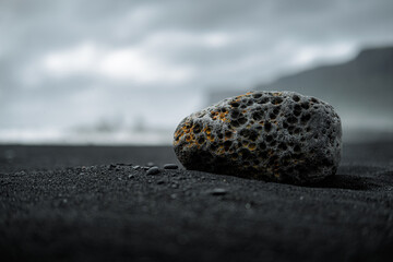 Close-up of a porous lava rock on a black sand beach under a moody, overcast sky. The dramatic lighting emphasizes the unique texture and volcanic origin of the stone. In the background, blurred sea s