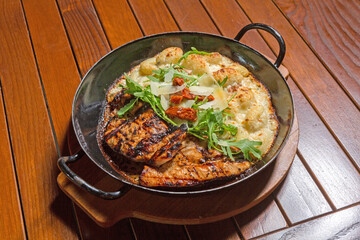 Grilled chicken breast served with creamy baked potatoes, arugula, sun-dried tomatoes, and parmesan in a black pan on a wooden table