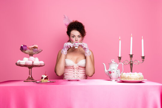 A modern woman embraces a whimsical baroque style with elegance, sipping tea at a pink table surrounded by vintage pastries