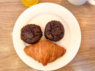 Two chocolate muffins and a croissant arranged on a white plate resemble a smiling face, adding whimsy to a cozy breakfast scene with drinks on the side.