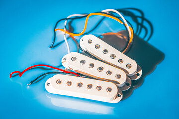 Three white electric guitar pickups with metal pole pieces and colorful wires are arranged on glossy blue surface, casting shadows. Components and wiring are clearly visible in the bright lighting