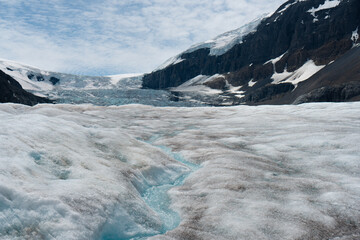 Glacial stream flowing across a vast, icy landscape beneath rocky mountains.