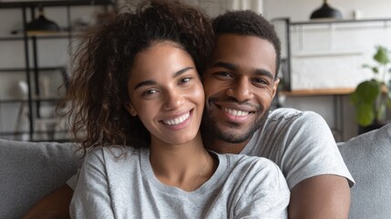 portrait of multiethnic couple embracing and looking at camera sitting on sofa smiling african american woman hugging mid adult man sitting on couch from behind at home happy mixed race couple laugh 