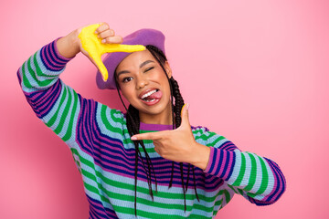 Playful young woman wearing a colorful sweater and beret making humorous hand gestures on a vibrant pink background