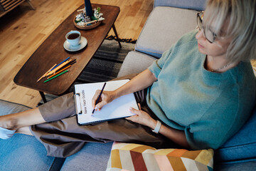Middle aged woman writing New year's goals on note paper. New year resolutions list, cup of tea on table. Goals, resolutions, plans, action, checklist concept. New Year template, copy space