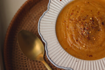 Creamy pumpkin soup in a rustic bowl on a woven tray, topped with chili flakes and pepper for a warm, cozy autumn meal