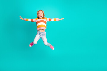 Obraz premium Smiling young girl joyfully jumping in a colorful outfit against a vibrant turquoise background.