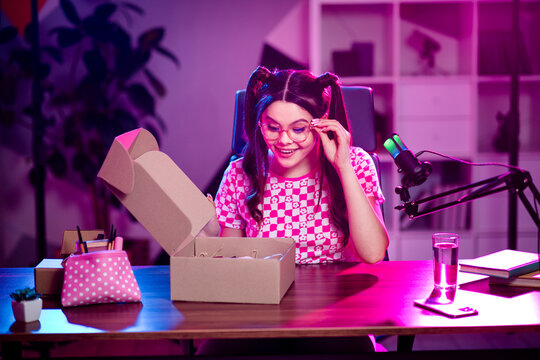 Popular female streamer in stylish setup unboxing package during live broadcast, illuminated by vibrant pink neon lighting - Powered by Adobe