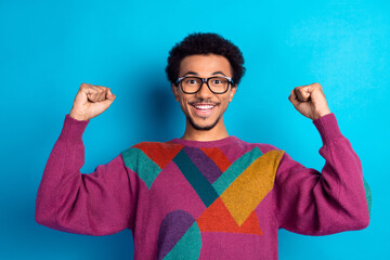 Cheerful young man in colorful retro sweater celebrates success with raised fists against a bright...