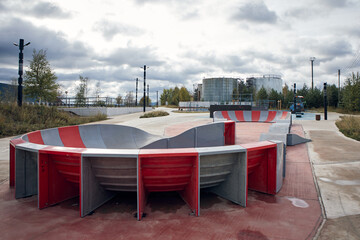 Modern skatepark with red and gray concrete ramps and curved design. Outdoor urban sports area under cloudy autumn sky. Contemporary architecture for youth recreation and skateboarding.