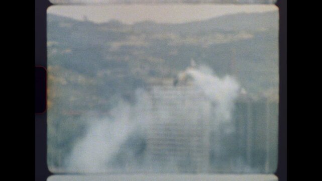 Archival footage in 8mm format depicting the controlled implosion of a towering structure that collapses inward, sending a billowing cloud of dust and debris into the air amidst a blurred cityscape