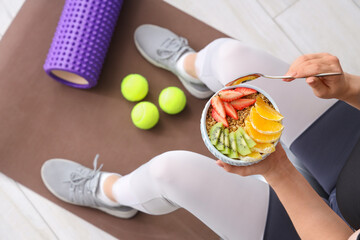 Sporty young woman sitting on yoga mat with bowl of nutritious oatmeal and sports equipment at...