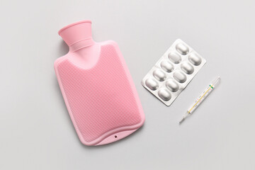 Hot water bottle with pills and thermometer on grey background