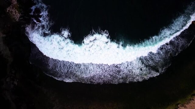 aerial view of ocean waves crashing against a dark, rocky coastline. The contrast of the white foam against the deep blue sea creates a beautiful and dramatic pattern in Gunung Kidul on April 28, 2024