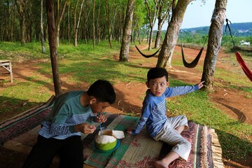 Two young boys eating from a coconut while sitting on a mat in a rural outdoor setting with trees and hammocks.