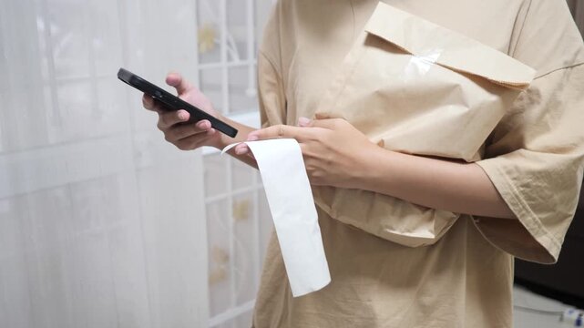 A woman holding a paper package and checking an online receipt using a smartphone. Concept of online shopping, e-commerce, digital payment, and parcel delivery at home. Modern lifestyle and technology
