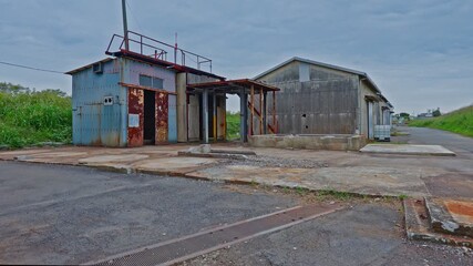 Two dilapidated, rustic industrial buildings with rusted metal siding and peeling paint stand on a cracked concrete pad in a rural or neglected area. - Powered by Adobe