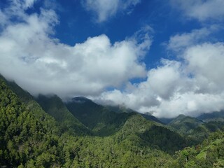 Obraz premium Aerial view of a lush green mountain covered with dense tropical forest under a bright blue sky and low clouds.