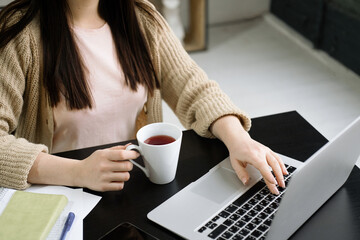 Close-up of woman's hands holding a cup of coffee while working on a laptop.